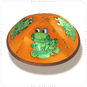 May include: Orange kippah featuring a cartoon frog design. The kippah has a brown trim and is decorated with multiple green frogs on a lily pad. The kippah is a traditional Jewish head covering.