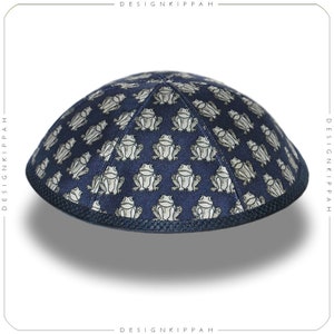 May include: A navy blue kippah featuring a repeating pattern of light gray frog illustrations. The kippah has a rounded shape with a dark blue trim. The text "DESIGN KIPPAH" is printed vertically on the left and right sides.