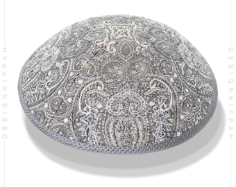 Puede incluir: Una kipá gris con un patrón de paisley blanco y dorado. La kipá tiene un borde gris.