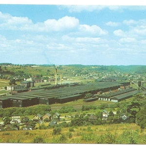 May include: Vintage postcard featuring an aerial view of a large industrial complex, likely a factory or manufacturing plant. The buildings are dark-colored with long, rectangular shapes. The sky is blue with scattered clouds, and the surrounding landscape is green with rolling hills.