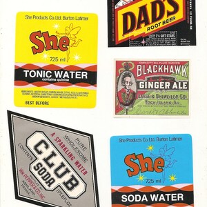 May include: A collection of vintage soda labels, including "Dad's Root Beer," "Blackhawk Ginger Ale," and labels for "She" brand tonic and soda water. The labels feature colorful designs and text, showcasing classic beverage branding.
