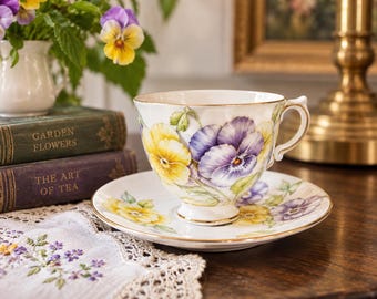 May include: A white porcelain teacup and saucer set featuring a delicate hand-painted pattern of yellow and purple pansy flowers. The rim of the cup and saucer is accented with a thin gold trim, and the cup has a curved handle and a pedestal base.