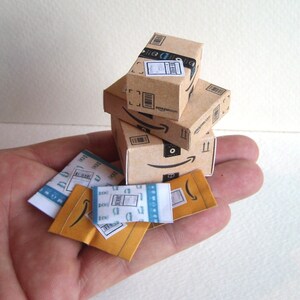 Peut inclure: Un ensemble de quatre petites bo&icirc;tes en carton avec des logos et des &eacute;tiquettes Amazon. Les bo&icirc;tes sont de diff&eacute;rentes tailles et sont empil&eacute;es les unes sur les autres. Il y a aussi trois enveloppes plus petites avec des logos et des &eacute;tiquettes Amazon.