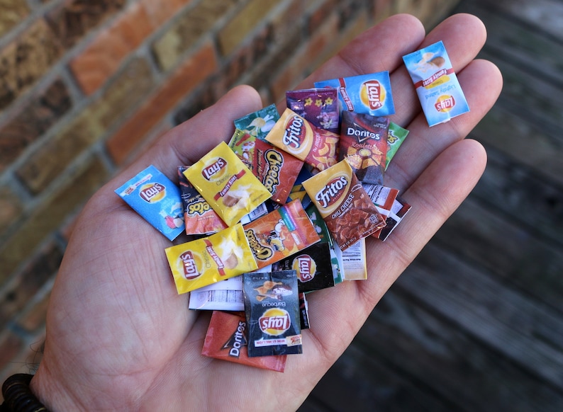 May include: A hand holding a variety of miniature snack chip bags. The bags are all different colors and feature various brand names, including Lay's, Fritos, and Doritos.