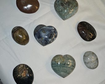 Op de afbeelding: Assortiment van gepolijste stenen in verschillende vormen, waaronder ovalen en hartvormige. De stenen vertonen een reeks kleuren, zoals bruin, groen en grijs, met unieke patronen en texturen. De collectie is gerangschikt op een wit oppervlak.