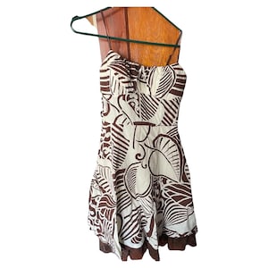 Puede incluir: Vestido sin tirantes, hasta la rodilla, con un estampado de hojas marrón y crema. El vestido está colgado de una percha verde. El dobladillo inferior tiene un ribete marrón.