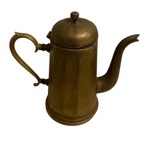 May include: A vintage brass coffee pot with a lid and a curved handle. The pot has a spout and a cylindrical body with vertical ridges. The handle is heart-shaped. The pot is made of brass and has a warm, golden color.