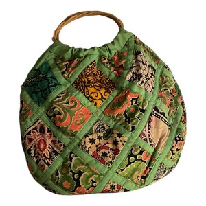 Puede incluir: Un bolso patchwork con asa tejida. El bolso presenta una variedad de cuadrados de tela estampada en tonos verdes, naranjas, amarillos y negros, cosidos con bordes verdes. El asa es un diseño tejido circular.