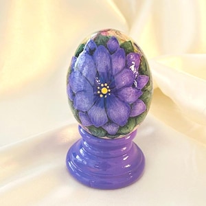 May include: A decorative egg featuring a hand-painted purple flower with a yellow center and green leaves, finished with a glossy sheen. The egg rests securely on a solid, smooth, circular purple pedestal stand with a tiered base design.