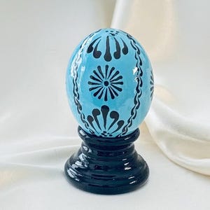 May include: A light blue egg-shaped ornament featuring intricate black hand-painted floral and geometric patterns. The egg rests securely on a glossy, solid black circular pedestal base, creating a striking contrast between the colors.