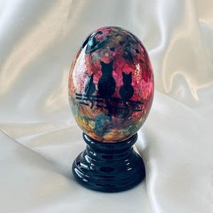 May include: An egg-shaped ornament featuring a colorful, marbled background of red, orange, yellow, and blue hues. The surface displays black silhouettes of three cats sitting on a fence. The ornament rests on a glossy, black, circular pedestal base.