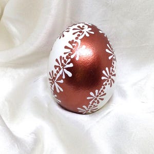 May include: A decorative egg featuring a metallic copper finish on one side and a white section on the other. A vertical band of white hand-painted floral patterns consisting of small petals and dots separates the two colors on the egg surface.