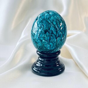 May include: A smooth, egg-shaped object with a vibrant teal and turquoise surface featuring a dark, intricate, branching crackle pattern. The object is securely mounted on a glossy, solid black, circular pedestal base with a tiered design.