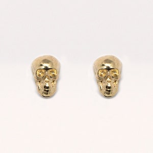 May include: A pair of gold skull stud earrings. The skulls are detailed with eye sockets and a nose.