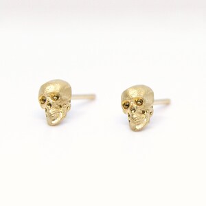 May include: A pair of gold skull stud earrings. The skulls are detailed and have small black eyes.
