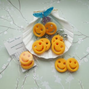 May include: A collection of yellow smiley face earrings displayed on a seashell. Some earrings are packaged in a clear bag with a white tag that reads "Vienna Sugar" and a website address. A blue butterfly decoration is also present.
