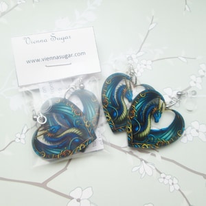 May include: Heart-shaped earrings featuring a blue dragon design with gold accents. The earrings are on silver-colored hooks. The image also includes a business card with the text "Vienna Sugar" and the website address.
