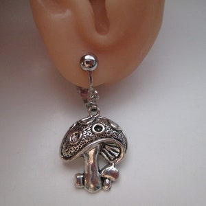 May include: A silver-colored mushroom-shaped earring. The earring is attached to a silver ball stud. The mushroom has a detailed design on the cap and stem. The earring is shown against a light skin tone.
