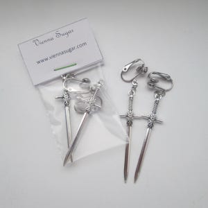 May include: A pair of silver sword-shaped clip-on earrings. The earrings are packaged in a clear plastic bag with a white card that reads "Vienna Sugar" and "www.viennasugar.com".