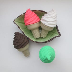 May include: Four miniature ice cream cones in different colours: pink, white, brown, and green. The cones are sitting on a green leaf-shaped dish and a white surface.