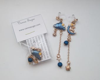 May include: A pair of gold dangle earrings with blue and gold charms. The charms include a cloud, a crescent moon, a star, and a bead. The earrings are packaged in a clear plastic bag with a white card that says "Vienna Sugar" and "www.viennasugar.com".