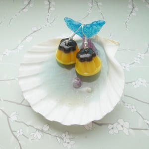 May include: A pair of novelty earrings shaped like yellow and brown pudding desserts. The earrings are suspended from silver hooks and rest on a white seashell-shaped dish. A blue, translucent, wing-like decoration is behind the earrings.