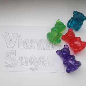 May include: Four gummy bear shaped candies in different colors: green, blue, red, and purple. The candies are arranged on a white surface with the words "Vienna Sugar" written in pencil.