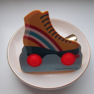 May include: A decorative pin shaped like a retro roller skate. The skate is yellow with red, blue, and white stripes, black laces, and red wheels. The pin is on a white plate with a gold rim. Text on the pin reads "Geddes.04/2021 Wenzhou China."