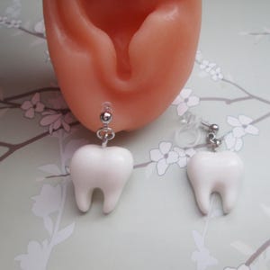 May include: White tooth-shaped earrings with silver-colored metal findings. The earrings are dangling and have a glossy finish. The background is a light green color with a floral pattern. The earrings are a fun and unique accessory.