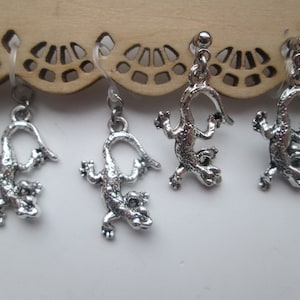May include: Four silver lizard earrings with a dangling design. The earrings are on a wooden background with a decorative pattern.
