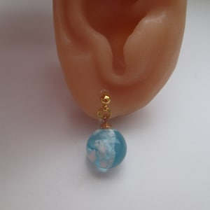 May include: A gold earring with a blue and white glass globe charm. The globe has white clouds inside.