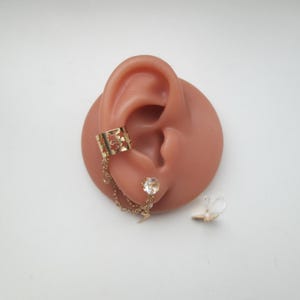 May include: Gold-toned ear cuff with a star-shaped cutout design, connected by a chain to a clear gemstone stud earring. A small, clear earring back is also present. The cuff is designed to wrap around the ear.