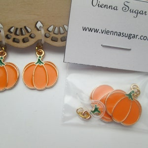 May include: Two orange pumpkin charms with gold-tone metal outlines and green stems, each attached to a clear plastic loop. A small plastic bag contains additional identical pumpkin charms, accompanied by a card labeled Vienna Sugar.