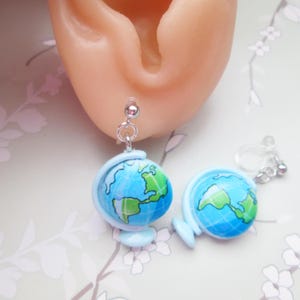 May include: A pair of novelty earrings featuring miniature globes. The globes are light blue with green and blue continents. The earrings have silver-colored metal findings and are attached to a person's ear.