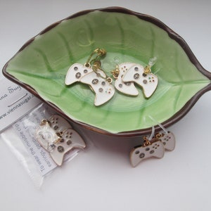 May include: Four pairs of white and gold video game controller earrings. The earrings are displayed on a green leaf-shaped dish. The text "Vienna Sug" and "www.viennasugar.com" is visible on a white card.