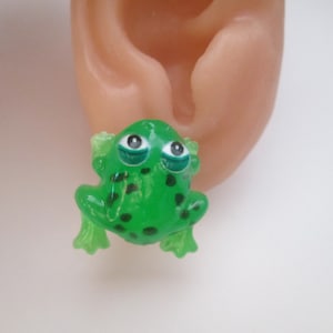 May include: A green frog-shaped earring with black spots and large, expressive eyes. The frog has a glossy finish and is positioned near a person's ear, suggesting it's being worn as jewelry. The earring is a whimsical accessory.
