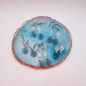 May include: A set of four pairs of dangle earrings featuring black rose cutouts and clear crystal accents. The earrings are displayed on a blue and gold agate-style plate.