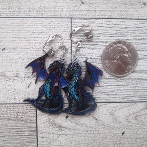 May include: A pair of clip-on earrings featuring a blue and purple dragon design. The earrings are approximately the size of a US quarter.