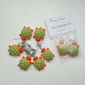 May include: Six pairs of green and orange turtle shaped clip-on earrings. The earrings are made of polymer clay and have a glossy finish. The earrings are packaged in a clear plastic bag with a white card that says "Vienna Sugar" and "www.viennasugar.com".