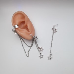 May include: Silver ear cuff and dangling star earrings. The ear cuff wraps around the ear with a chain that dangles down. The earrings feature small star charms hanging from delicate chains. The jewelry is displayed on a light-colored background.
