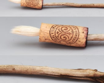 May include: A handmade brush with a driftwood handle, leather accents, and a wine cork ferrule. The brush has a goat hair tip and measures 11.2 inches overall. The cork is stamped with a design.