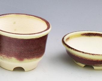 May include: A ceramic bonsai pot and a matching shallow saucer. Both pieces feature a cream-colored interior and rim, with a textured, deep reddish-brown glaze covering the exterior. Each item has a small footed base with a cutout arch detail.
