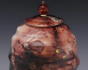 May include: A handcrafted ceramic jar with a lid. The jar features a textured surface with shades of brown, black, and white. The lid is topped with a small, round, amber-colored knob. A unique decorative piece.