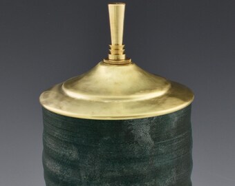 May include: A handcrafted ceramic vessel with a dark teal, textured exterior and a gold-colored metal lid and finial. The jar has a cylindrical shape with a slightly flared base. The lid is a flat disc shape.