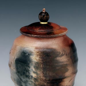 May include: A handcrafted ceramic jar with a lid. The jar has a rounded shape with a textured surface. The colors are earthy, with shades of brown, black, and tan. The lid is a darker brown with a small, decorative knob on top.