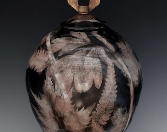 May include: A black ceramic vase with a lid and a decorative butterfly-shaped handle. The vase features a pattern of light brown and white leaves and ferns. The vase has a rounded body and a narrow base.