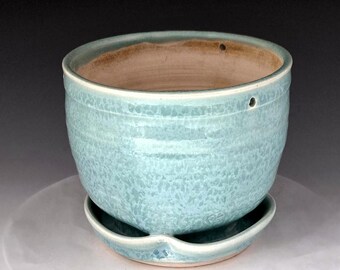May include: A handcrafted, light blue ceramic planter with a matching saucer. The planter has a textured surface and a wide opening. The interior is a contrasting tan color. Two small drainage holes are visible near the top rim. The saucer has a curved design.