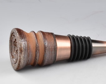 May include: A decorative wine bottle stopper with a rustic design. The stopper features a brown, textured ceramic top, a copper-colored metal band, a black rubber seal, and a pointed metal tip. The design is elegant and suitable for wine enthusiasts.