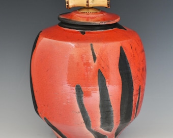 May include: A ceramic jar with a vibrant red glaze and bold black vertical brushstroke patterns. The jar features a matching red lid topped with a light-colored, textured bamboo-shaped handle. A small maker's mark is visible near the base.