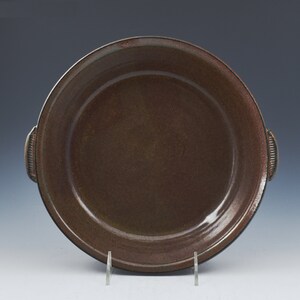 May include: A round, brown ceramic serving dish with two small handles on either side. The dish has a slightly raised rim and a smooth, dark brown interior. The handles have a textured, ribbed design. The dish is displayed on a clear stand.